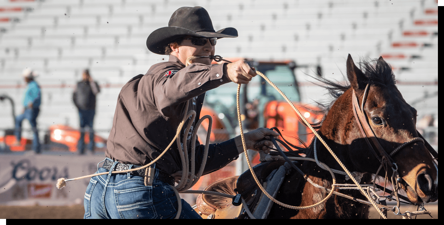 Chet Weitz wearing a black cowboy hat, brown shirt, and blue jeans dismounting his horse with a rope in his mouth. 